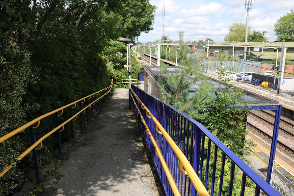 A narrow elevated walkway with a gravel surface, bordered by a yellow and blue metal railing, runs alongside a green, bushy area with dense foliage on the left. On the right, there is a view of a train station platform with yellow safety lines along the tracks, and an adjacent parking lot filled with white and blue cars. The background shows an overcast sky with scattered clouds, mature trees, shrubs, and ongoing landscaping or maintenance work, consistent with garden care and landscaping themes for Harold Wood station front gardens, as managed by Gardeners Gallows Corner.