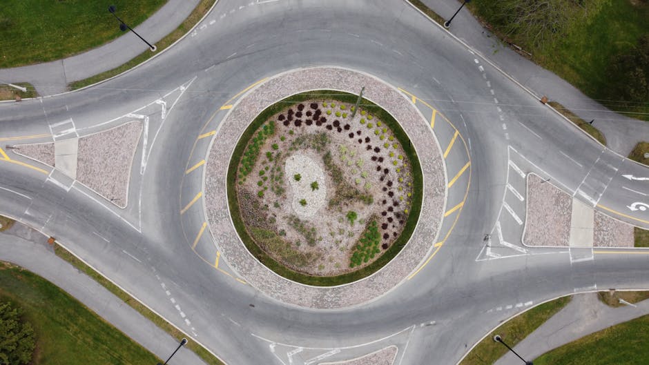 An aerial view of Gallows Corner roundabout showing a landscaped central island with a variety of plants and shrubs arranged in a circular pattern. Surrounding the island, the road features clear white markings, including directional arrows and lane indicators. The grass verges and paved footpaths are visible along the edges, with a neatly trimmed hedge lining part of the perimeter. The central planting area includes a mix of small bushes, ornamental grasses, and flowering plants, some with purple, green, and brown foliage, creating a visually appealing contrast against the gravel and soil surface. The overall scene showcases well-maintained garden beds integrated into the roundabout’s layout, emphasizing landscaping work that enhances the traffic roundabout while ensuring ease of maintenance, with no visible tools or ongoing gardening activities. The lighting is natural, indicating daytime with soft shadows, and the setting reflects professional garden maintenance by Gardeners Gallows Corner.