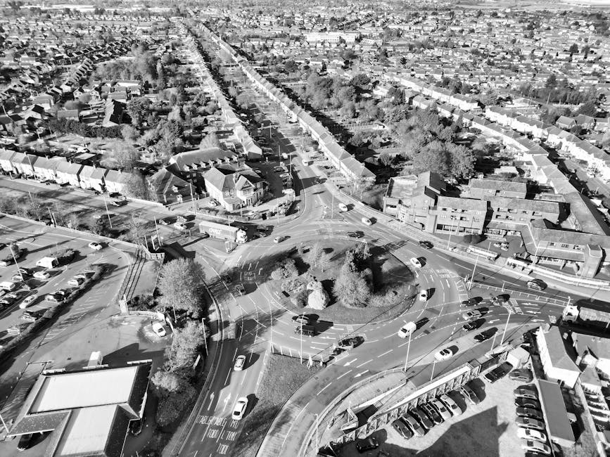 An aerial view of Gallows Corner roundabout showing a landscaped central island with a variety of plants and shrubs arranged in a circular pattern. Surrounding the island, the road features clear white markings, including directional arrows and lane indicators. The grass verges and paved footpaths are visible along the edges, with a neatly trimmed hedge lining part of the perimeter. The central planting area includes a mix of small bushes, ornamental grasses, and flowering plants, some with purple, green, and brown foliage, creating a visually appealing contrast against the gravel and soil surface. The overall scene showcases well-maintained garden beds integrated into the roundabout’s layout, emphasizing landscaping work that enhances the traffic roundabout while ensuring ease of maintenance, with no visible tools or ongoing gardening activities. The lighting is natural, indicating daytime with soft shadows, and the setting reflects professional garden maintenance by Gardeners Gallows Corner.